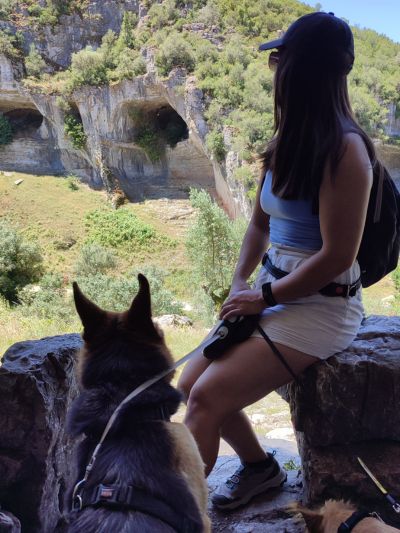 Mulher equipada para caminhada e sentada numa rocha a observar a paisagem com dois cães no chão perto dela presos por trelas. Trilhos pet friendly em Portugal - buracas do Camilo