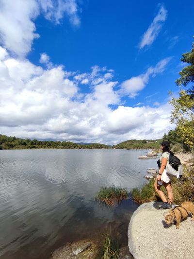 Mulher junto a um lago, olhando para a paisagem, com um cão pequeno de peitoral e um gato em mochila de transporte. Cenário natural com céu nublado e vegetação ao redor do lago. Um dos trilhos pet-friendly em portugal