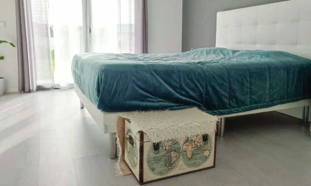 Quarto com baú decorativo e coberto por manta, colocado no chão encostado à cama - tem o objetivo de servir como degrau para melhorar a mobilidade do cão idoso dentro de casa