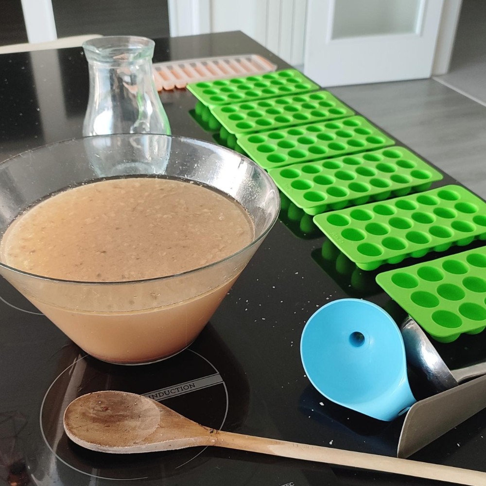 imagem de uma bancada de cozinha preta com diversos utensílios: cuvetes de gelo, colher de pau, jarro, funil pequeno, espátula, colher de servir e uma taça com caldo de ossos dentro de uma taça de vidro.